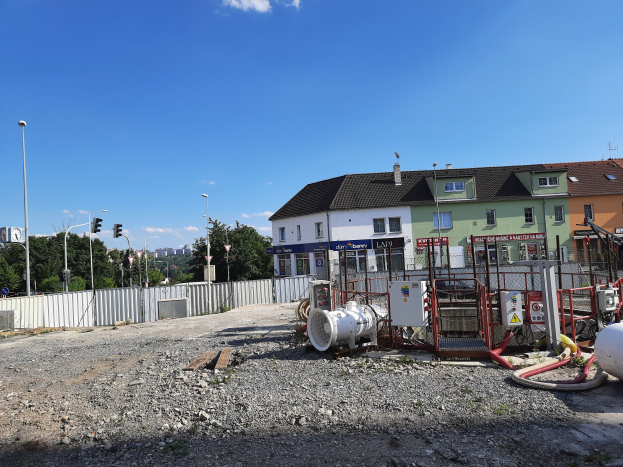Baustelle mit freiliegenden Rohren und Geräten auf dem Boden, umgeben von Gebäuden, Laternen, Ampeln, einem Zaun, Bäumen und einem bewölkten Himmel.