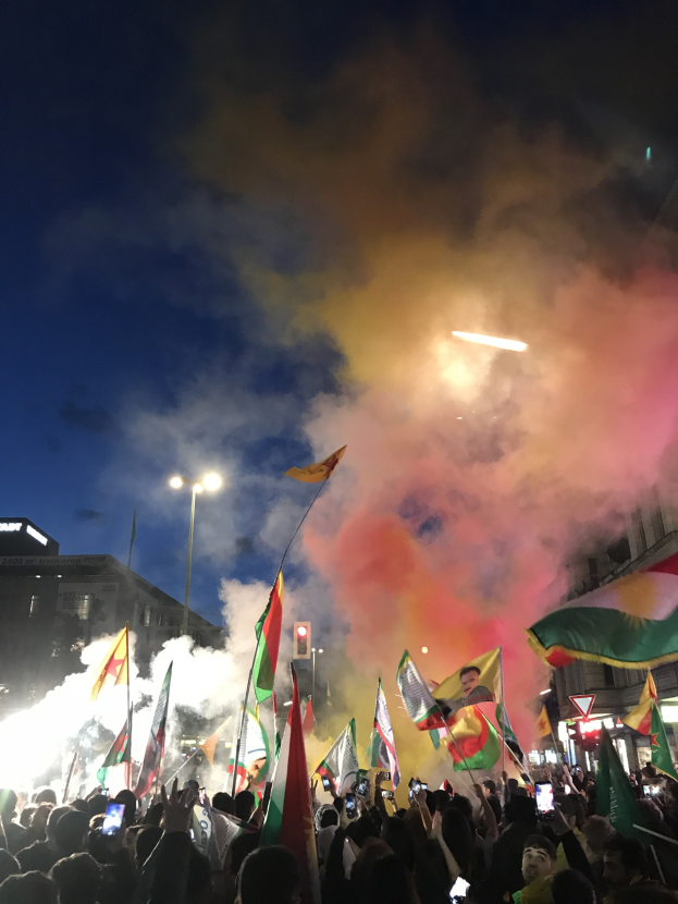 Eine Menschenmenge vor einem Gebäude mit Fahnen schwenkend und Handys haltend unter farbigen Feuerwerkskörpern im Nachthimmel.