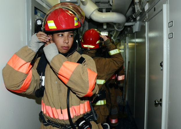 Feuerwehrleute in Uniform stehend in einem Raum während einer Übung, mit Rohren und Gegenständen im Hintergrund.