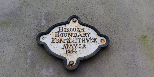 Metallschild an einem Gebäude mit schwarzem Hintergrund und weißer fetter Schrift, die den Text "Stadtteilgrenze Edm Smithwick Bürgermeister" zeigt.
