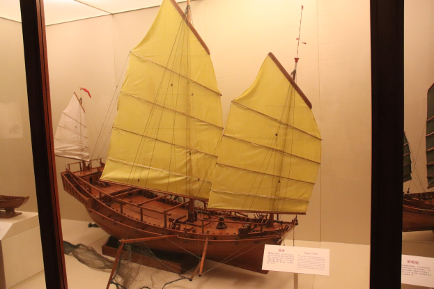 Ein hölzernes Modellsegelboot in einer Glasvitrine mit Informationsschildern und einer Wand im Hintergrund.