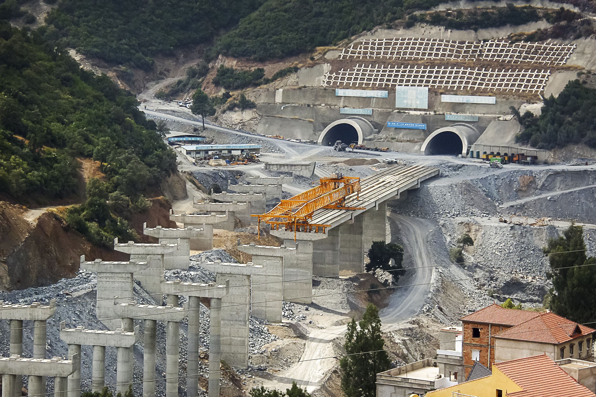 Großer Baustelle mit einem zentralen Tunnel, umgeben von Bäumen, Gebäuden, Fahrzeugen, Säulen, Felsen und einer Straße unten.