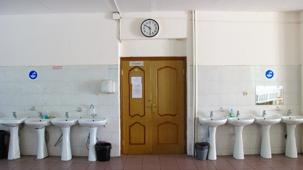 Ein Badezimmer mit mehreren Waschbecken, bodenebenen Mülltonnen, einer Tür im Hintergrund, einer Wanduhr und einem Spiegel auf der rechten Seite.
