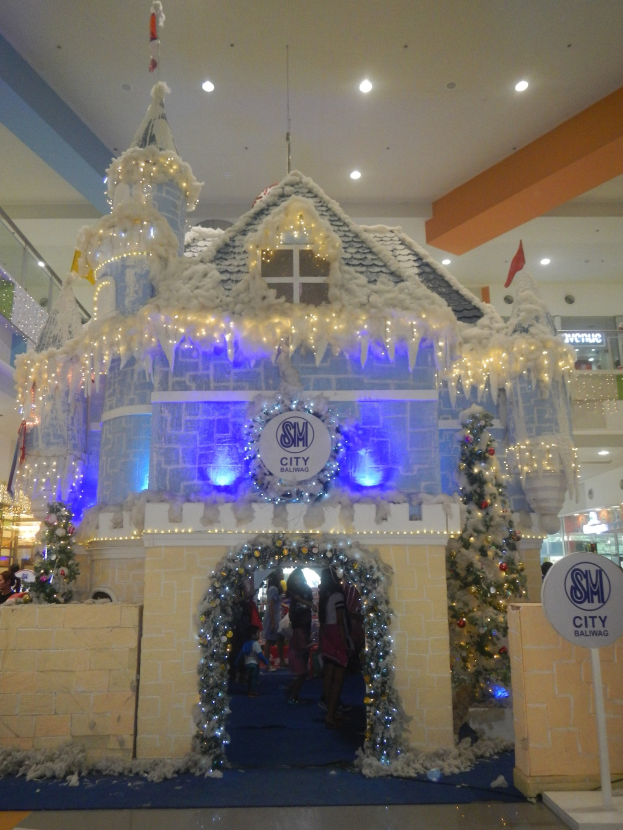 Ein festliches Weihnachtsdorf mit einem burgenähnlichen Aufbau mit Lichtern und Dekorationen, umgeben von Menschen in einem Einkaufszentrum mit beleuchteter Decke.