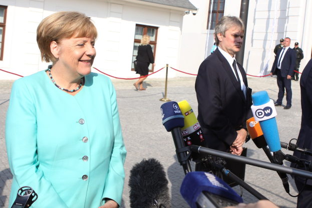 Bundeskanzlerin Angela Merkel bei einer Pressekonferenz vor einem Gebäude mit Fenstern stehend, umgeben von Menschen mit Mikrofonen und einer Absperrung unter einem bewölkten Himmel.