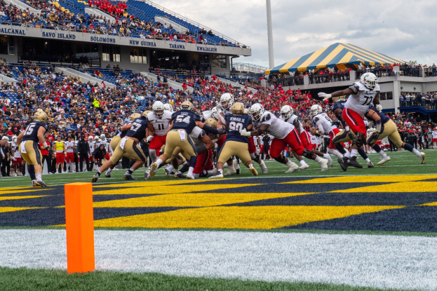 Fußballspieler in Helmen konkurrieren auf einem Rasenfeld, während Zuschauer auf Tribünen unter einem bewölkten Himmel sitzen, mit einem Zelt und anderen Gegenständen im Hintergrund.