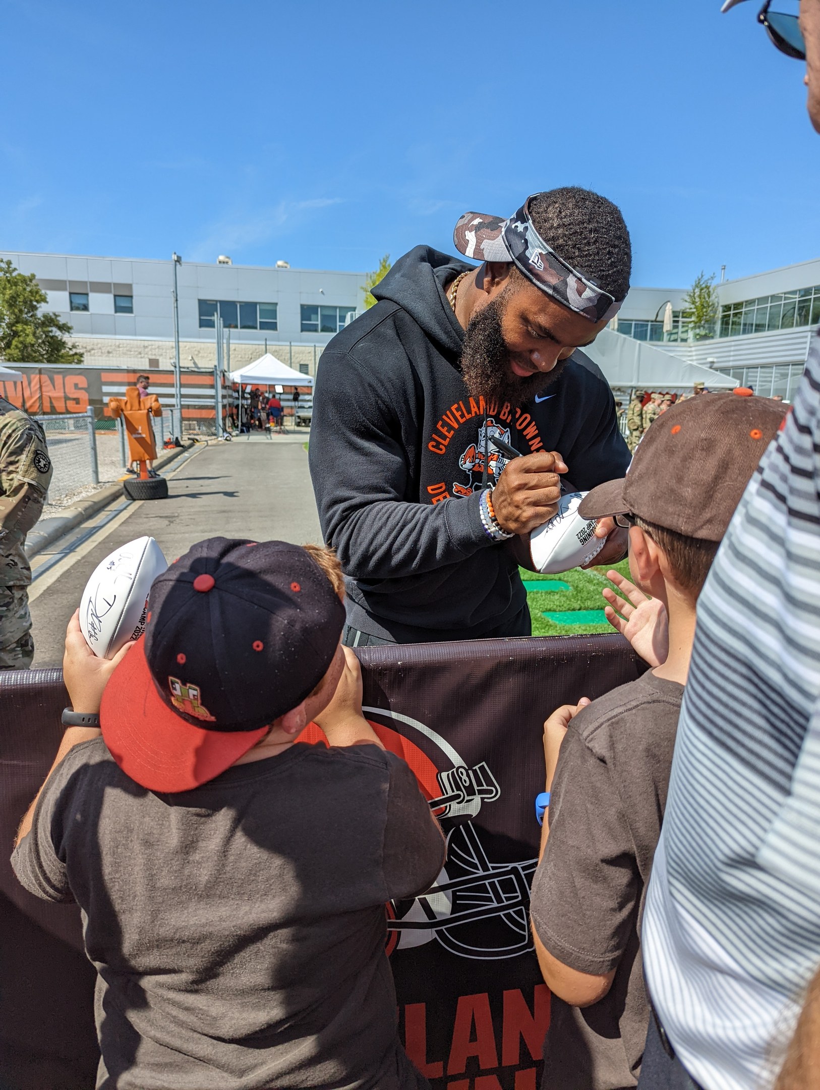 Cleveland Browns Wide Receiver Odell Beckham Jr. signiert Autogramme für Fans vor der Trainingsanlage des Teams, umgeben von Menschen mit Mützen, einem Banner, Gebäuden und Bäumen im Hintergrund unter einem bewölkten Himmel.