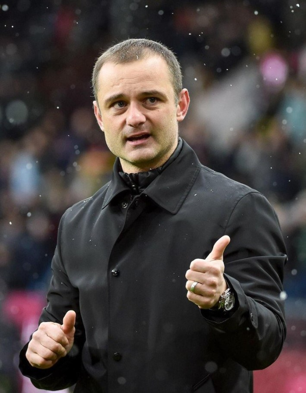 Ein Mann in einer schwarzen Jacke zeigt den Daumen hoch, trägt eine Uhr, mit einem unscharfen Hintergrund.