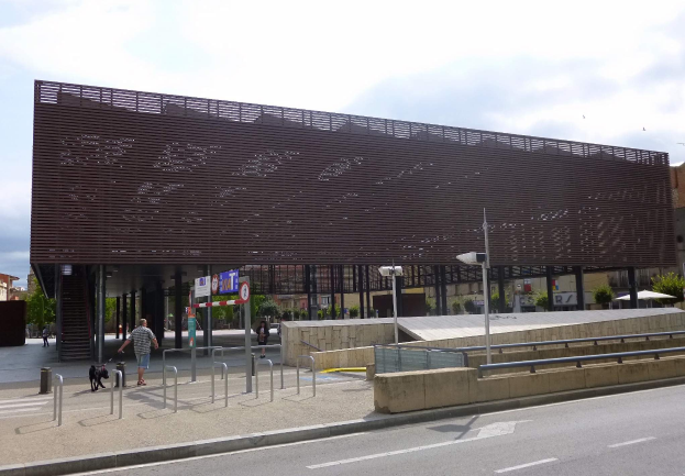 Gro├če modernes Geb├Ąude mit zahlreichen Fenstern, das als neuer Eingang ins Stadtzentrum dient, umgeben von Stra├čeninfrastruktur, Fahrzeugen, Passanten und B├Ąumen unter einem bew├Âlktem Himmel.