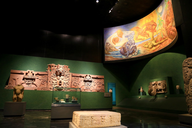 Ein Ausstellungsraum im Anthropologischen Museum von Mexiko-Stadt mit verschiedenen Artefakten, darunter Skulpturen, Tischgegenstände, eine Texttafel, eine Wandmalerei und Deckenbeleuchtung.