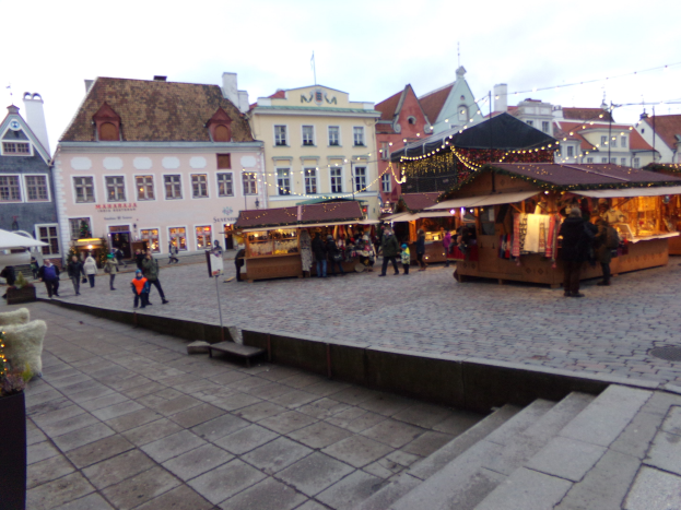 Ein geschäftiger Weihnachtsmarkt in Tallinn, Estland mit Menschen um dekorierte Stände, festliche Lichter, bewölkter Himmel, Stufen nach oben und Topfpflanzen im Bereich.