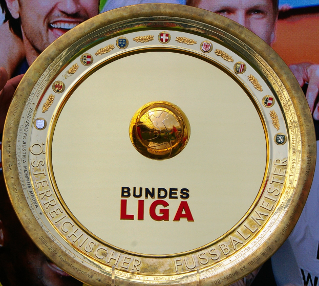 Ein Mann hält einen goldenen Schild mit der Aufschrift "Bundesliga" in die Höhe, im Hintergrund eine Fahne mit drei Personen.