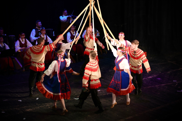 Eine Gruppe von Menschen in bunten Kostümen tanzt um einen Maibaum, einige halten Holzstöcke, während andere im Hintergrund Musikinstrumente spielen.