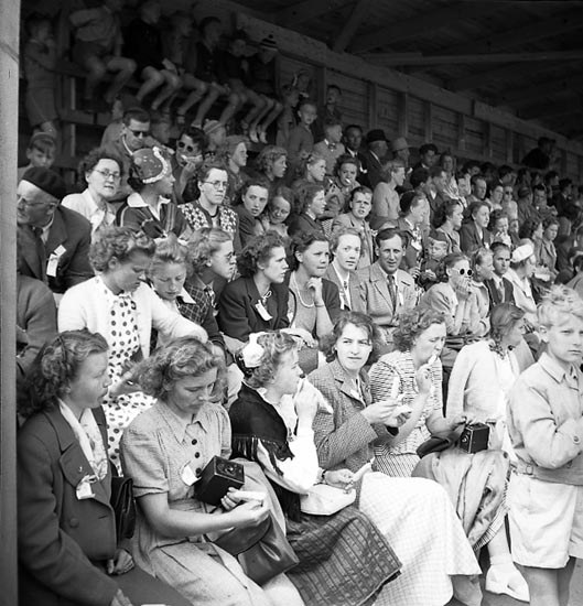 Schwarzes und weißes Foto einer Menschenmenge, die in einem Stadion mit einer hölzernen Wand und einem Dach im Hintergrund sitzt, wobei einige Kameras halten.