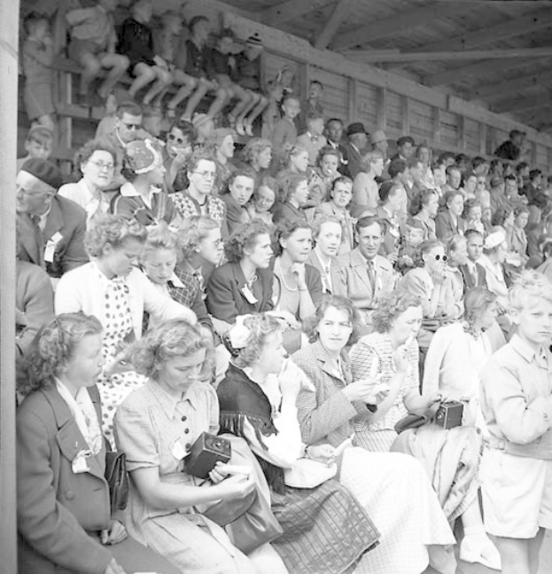 Schwarzes und weißes Foto einer Menschenmenge, die in einem Stadion mit einer hölzernen Wand und einem Dach im Hintergrund sitzt, wobei einige Kameras halten.