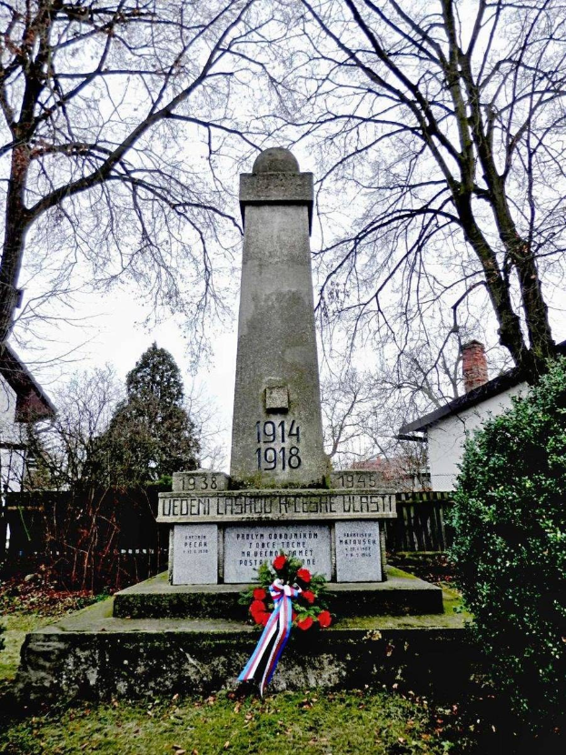 Ein Weltkriegsgedenkmal auf einem Friedhof, geschmückt mit einem Kränzchen, umgeben von Gras und welken Blättern, mit Pflanzen, Bäumen, Häusern und Himmel im Hintergrund.