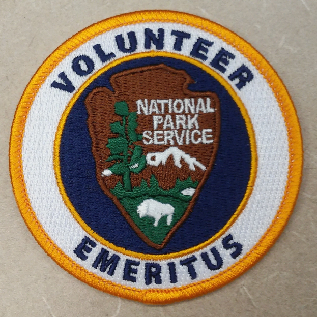Runder Aufnäher mit "Volunteer National Park Service Emeritus" in schwarzer Schrift auf einem weißen Hintergrund, umrandet in Blau mit einem gelben Kreis um den Text.