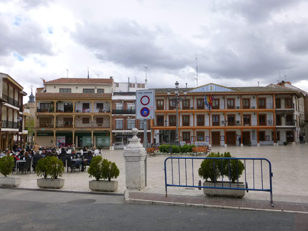 Ein belebter Stadtplatz mit sitzenden und stehenden Menschen, umgeben von Topfpflanzen, Metallabsperrungen, Schildern, Straßenmasten mit Flaggen, Gebäuden und einem bewölkten Himmel.