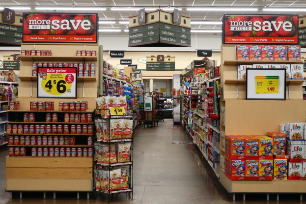 Ein Kiosk-Innenraum mit Regalen und Boden voller verschiedener Lebensmittel, beleuchtet von Deckenlampen, mit Informationsschildern und einer Person im Hintergrund.