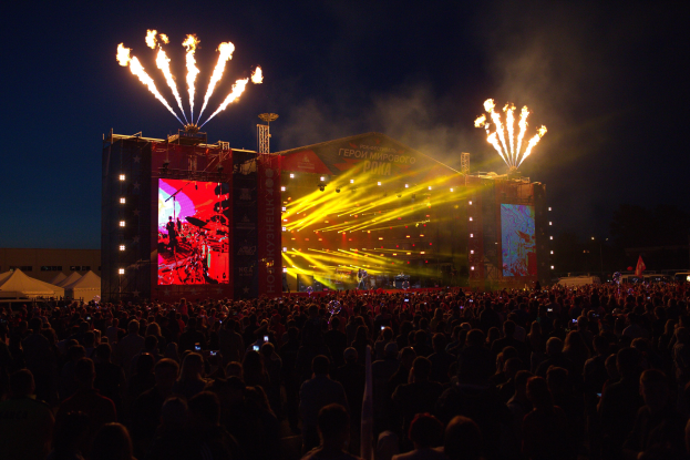 Eine große Menschenmenge versammelte sich auf einem Musikfestival, mit einer Bühne in der Mitte, auf der Musiker Instrumente spielten, beleuchtet von hellen Lichtern und umgeben von Bannern, Zelten links und einem dunklen Himmel mit Feuerwerk im Hintergrund.