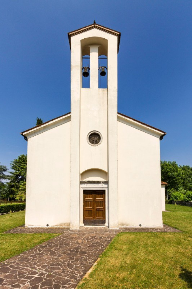Kleine weiße Kirche mit einem zentralen Glockenturm, umgeben von einem Weg, Gras, Pflanzen, Bäumen und einem bewölkten Himmel, wobei die Glocke laut läutet.