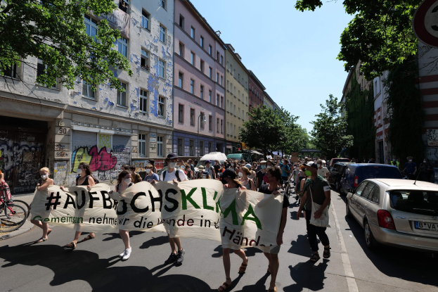 Eine Gruppe von Menschen marschiert auf einer Straße in Berlin, hält ein Transparent, mit einem Fahrradfahrer auf der linken Seite und parkenden Autos auf der rechten Seite, vor einem Hintergrund von Gebäuden, Bäumen und einem Schild.