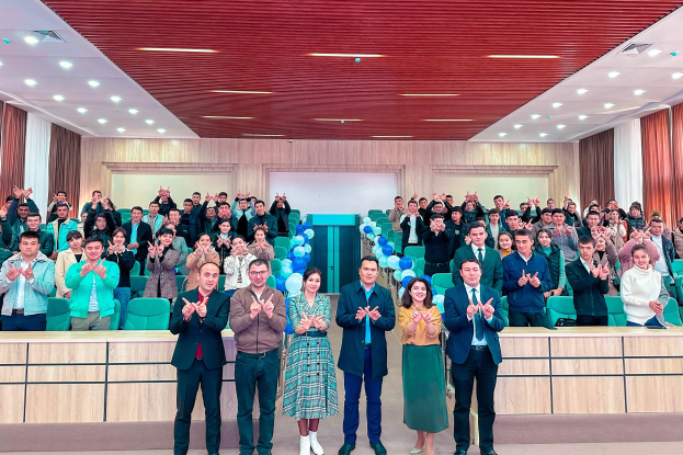 Eine Gruppe von Menschen steht vor einem Publikum, einige klatschen, in einem Raum mit Stühlen, Tischen, Ballons, Vorhängen und Deckenleuchten, bei der Eröffnung einer neuen Schule.