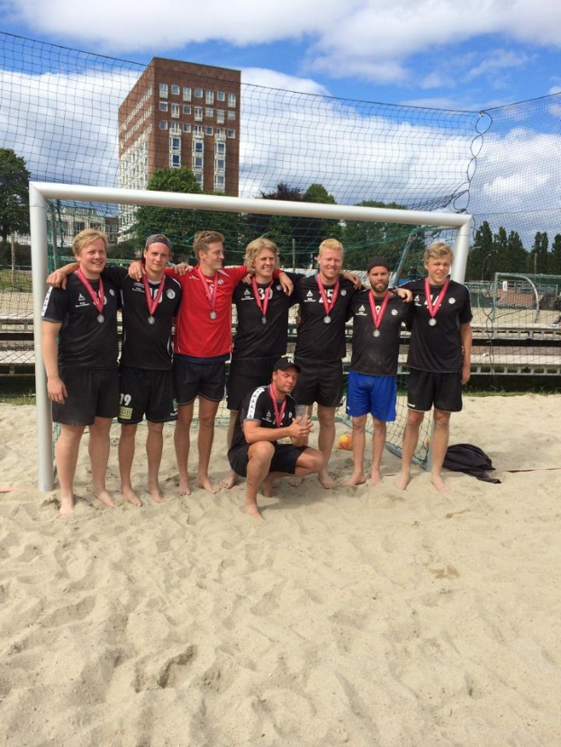 Mannergruppe in Sportkleidung auf einem sandigen Strand, mit Medaillen umgeben und von einem Netz, mit Bäumen, Gebäuden und einem bewölkten Himmel im Hintergrund.