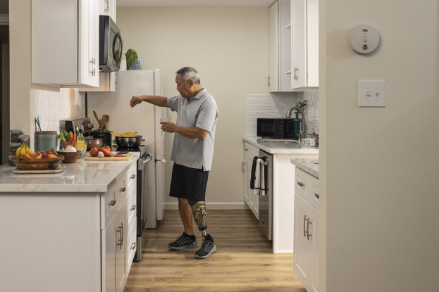 Ein Mann steht in einer Küche mit weißen Schränken und hält ein Glas in der Hand, während verschiedene Gegenstände wie Obst, Gemüse, Schüsseln und Löffel auf einer Arbeitsplatte links von ihm liegen.
