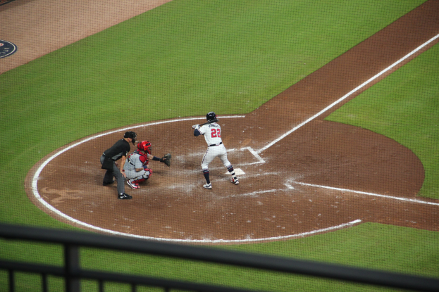 Baseballspieler in Uniform, der einen Schläger auf einem grünen Feld schwingt, mit einem Geländer am unteren Bildrand.