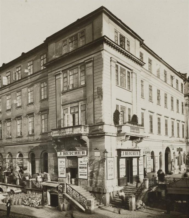 Altes Backsteingebäude mit der Aufschrift 'J Reicher Hotel' in Berlin, das Treppen mit Geländern, Blumenkästen mit Pflanzen und Informationsschildern sowie einige Menschen und Himmel im Hintergrund zeigt.