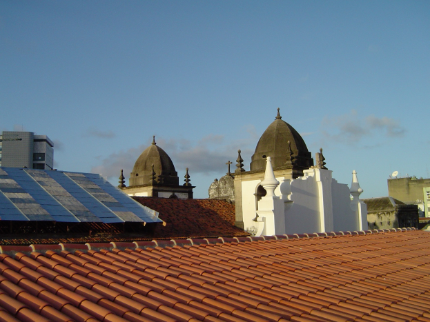 Stadtpanorama mit Gebäuden im Vordergrund, Solarpanelen auf einem Dach und einem klaren blauen Himmel im Hintergrund.