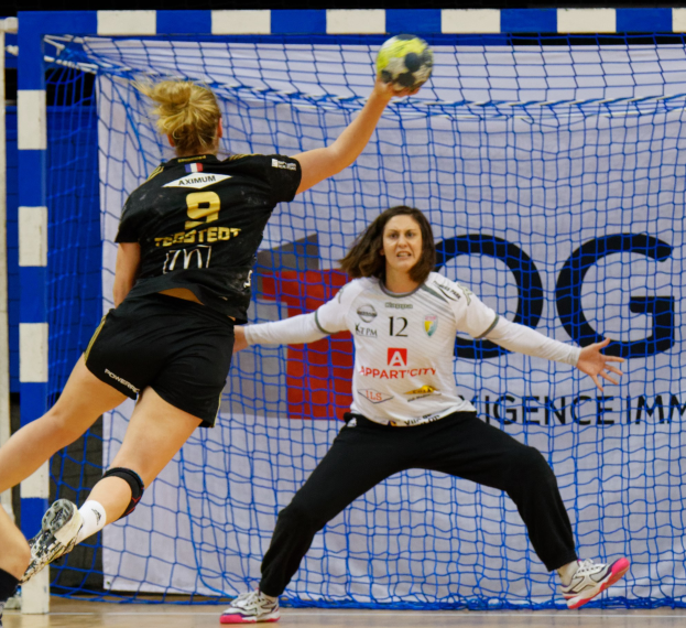 Zwei Frauen beim Handball vor einem Tor, eine hält den Ball, im Hintergrund ein Netz und eine Texttafel.