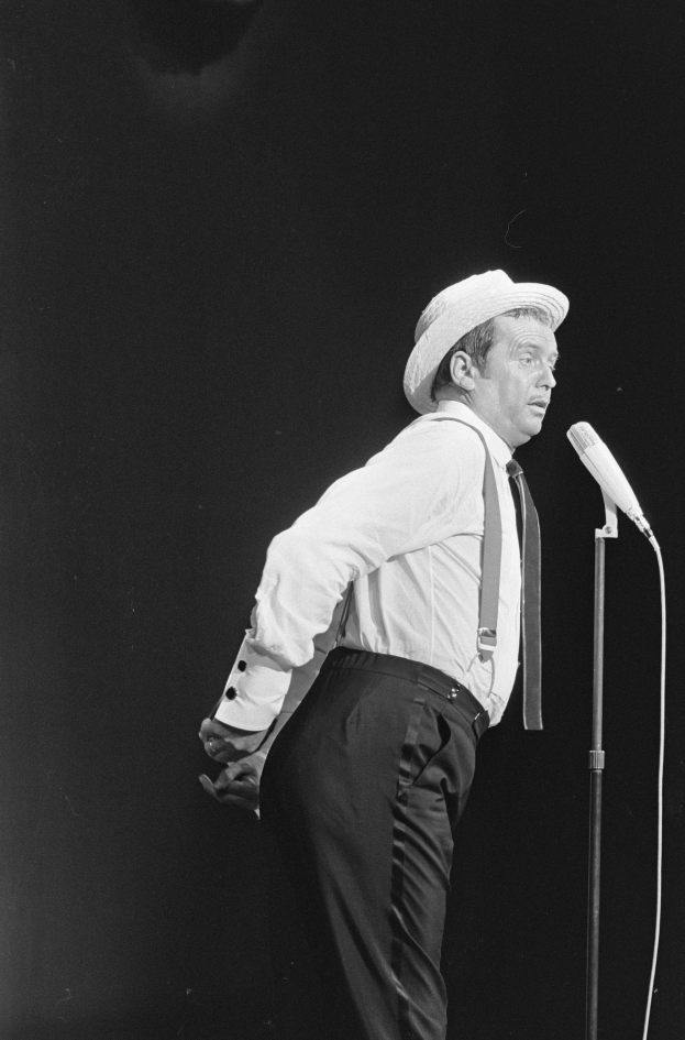 Ein Mann in einem weißen Hemd, schwarzen Hosen und Hut singt in ein Mikrofon auf einer Bühne.