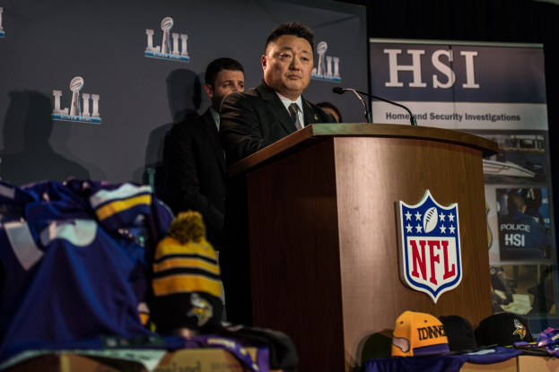 Ein Mann in einem schwarzen Blazer, wei├čem Hemd und schwarzer Krawatte spricht an einem Podium mit Mikrofon und Logo vor einer Menge. Blau-gelbes Merchandise ist links von ihm sichtbar, und ein "Super Bowl LII"-Banner ist im Hintergrund zu sehen.