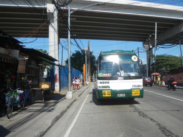 Ein grüner und weißer Bus fährt auf einer Straße mit Strommasten und -leitungen an Geschäften und Fußgängern vorbei, die eine Brücke überqueren, mit Fahrrädern, die an der Straße geparkt sind, und Bäumen im Hintergrund bei klarem Himmel.