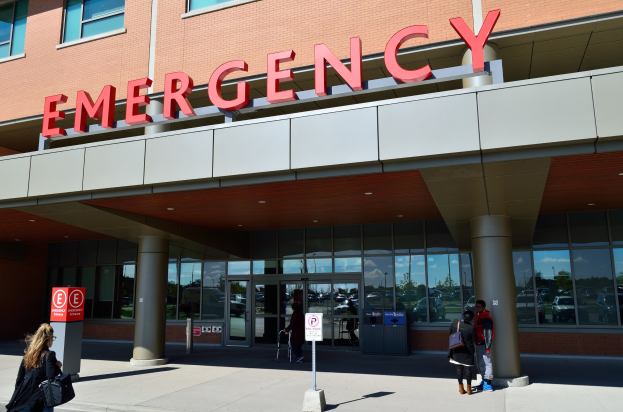 Eine Gruppe von Menschen geht vor einem Notfallgebäude mit Glaswänden und Säulen, einem Namensschild, Hinweistafeln auf Pfählen und einem Abfalleimer vorbei.