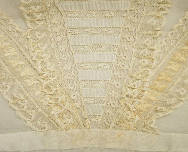 Nahaufnahme eines weißen Kleides mit zarter, kunstvoller Spitzenverzierung auf einem leichten, luftigen Stoff.