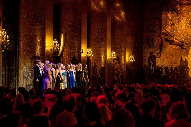 Eine Gruppe von Menschen auf einer beleuchteten Bühne im Royal Opera House in London mit bemalten Wänden, während das Publikum darunter zusieht.