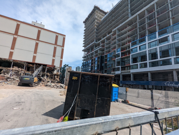 Großes Gebäude mit umfangreichem Schutt davor, umgeben von einem Metallzaun, umgeben von Baustoffen, Vegetation und Fahrzeugen unter einem bewölkten Himmel, was auf eine Baustelle für einen neuen Wohnkomplex hinweist.