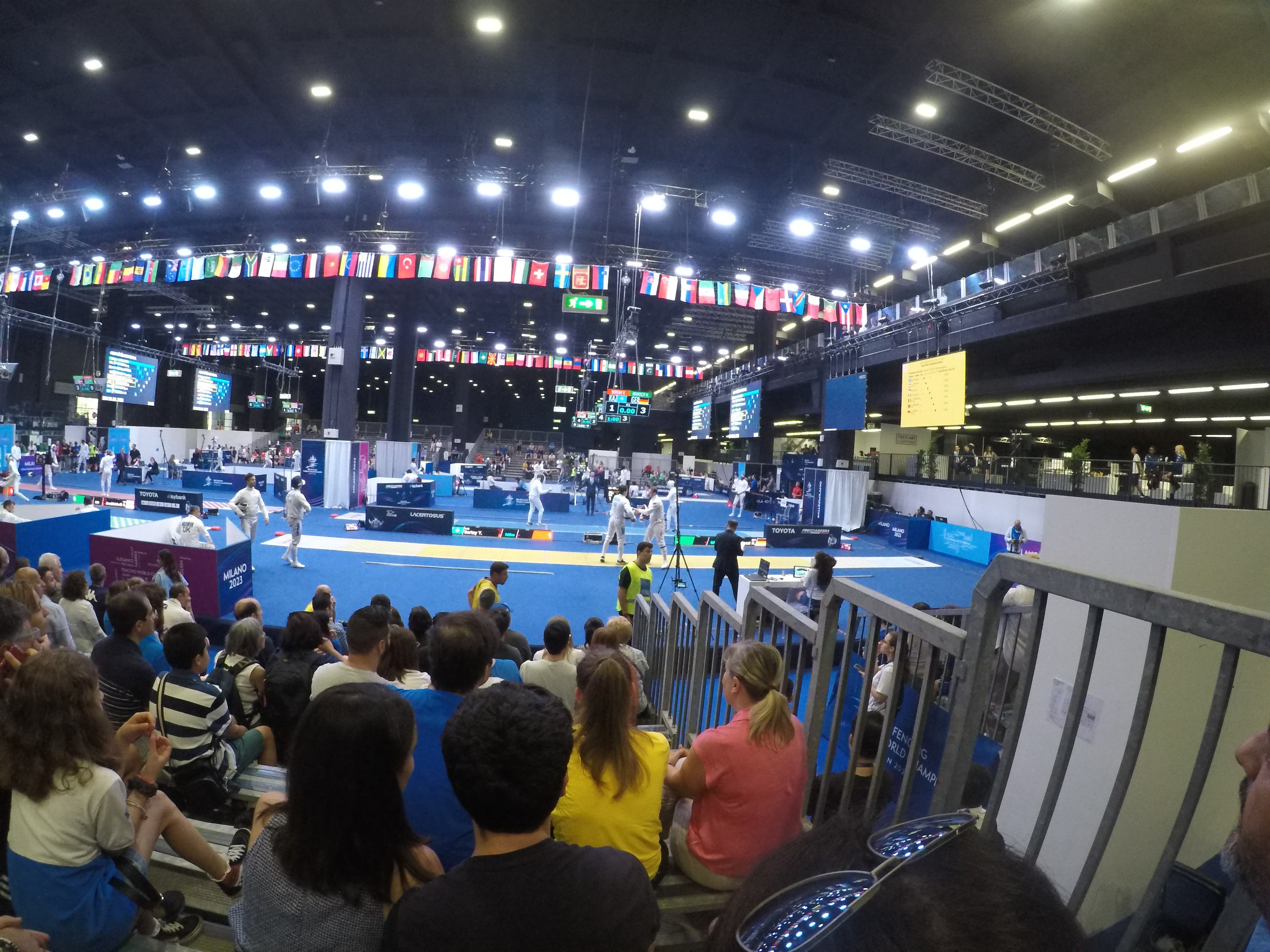Große Zuschauermenge sitzend und stehend in einer Indoor-Sportarena mit Bildschirmen, Fahnen und Deckenbeleuchtung im Hintergrund.