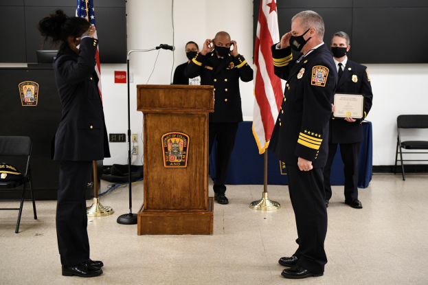Gruppe uniformierter Personen in Masken bei einer Vereidigung eines neuen Feuerwehrchefs, der eine Urkunde hält, mit Fahnen, Podium, Stühlen, Tischen und einer Wand im Hintergrund.