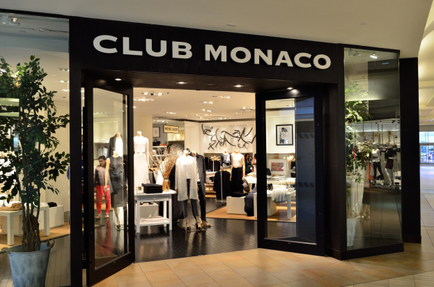 Vitrine eines Club Monaco Einzelhandelsgeschäfts in einem Einkaufszentrum, mit Glaswänden und -türen, Schaufensterpuppen in Kleidern, Auslagetischen mit Waren, einem Topf mit einer Pflanze und Deckenbeleuchtung.