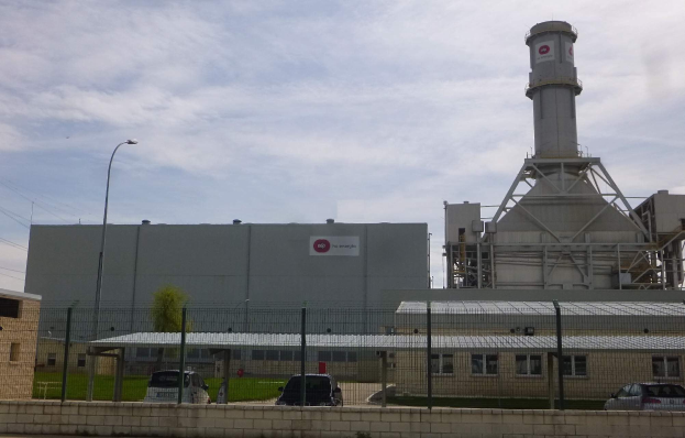 Großes industrielles Gebäude mit einem zentralen Turm, umgeben von einem Zaun, mit Fahrzeugen auf der Straße davor, Polen, Lampen, Drähte, Bäume, Gras und einem bewölkten Himmel im Hintergrund, Teil einer neuen Energiespeicheranlage.