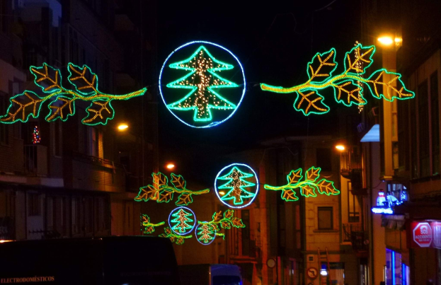 Eine nächtliche Stadtstraße mit festlichen Weihnachtslichtern in Form von Blättern, mit Fahrzeugen auf der Straße und Gebäuden auf beiden Seiten.