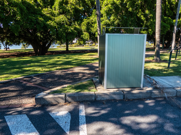 Transportable Toilette am Straßenrand in einem Park, umgeben von Bäumen und Gras mit einem Gewässer und einem klaren blauen Himmel im Hintergrund.