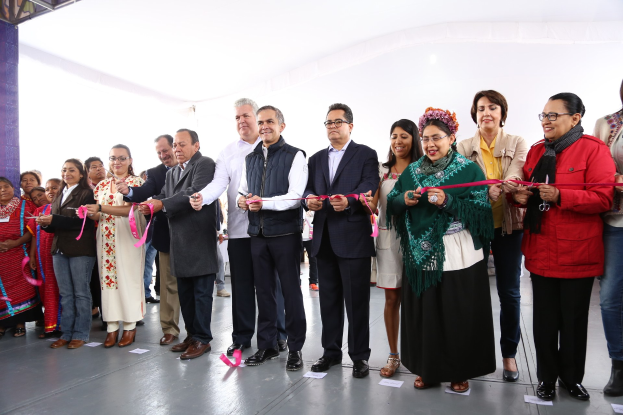 Eine Gruppe von Menschen, die strahlend eine Schere halten und vor einer Menge stehen, mit einer Säule links daneben, verstreuten Papieren auf dem Boden und einem weißen Tuch im Hintergrund.