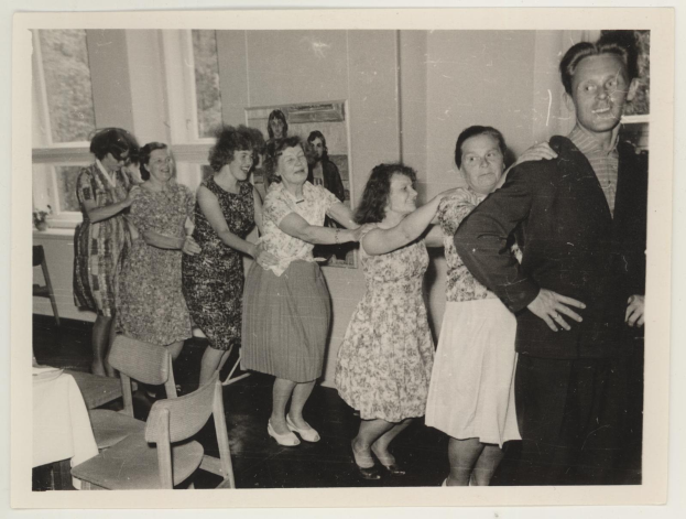 Eine Gruppe von Menschen, die in einem Raum im 1950er-Stil tanzen, mit Stühlen, einem Tisch mit einer wei├čen Decke, einem Foto auf der Wand und Fenstern im Hintergrund.