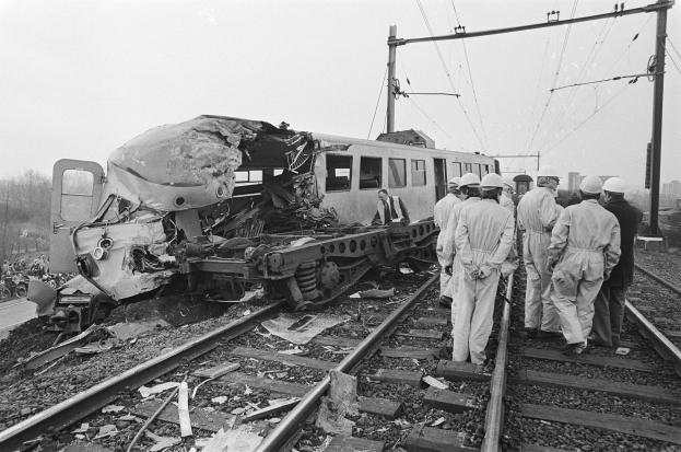 Schwarzes und weißes Bild einer Zugkollision auf den Gleisen mit Einsatzkräften in Helmen und naher Infrastruktur einschließlich Strommasten, Drähten, Bäumen, Gebäuden und einem klaren Himmel.