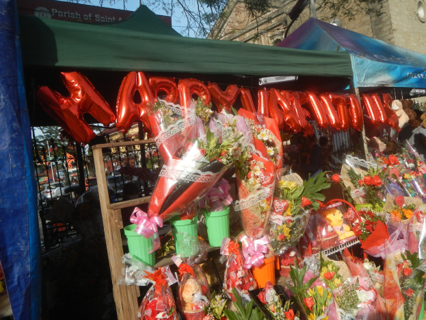 Ein Blumenladen, der für Valentinstag mit Sträußen, Teddybären und einem "Happy Valentine's Day"-Schild dekoriert ist, vor einem Hintergrund aus Gebäuden, Bäumen und einem klaren blauen Himmel.